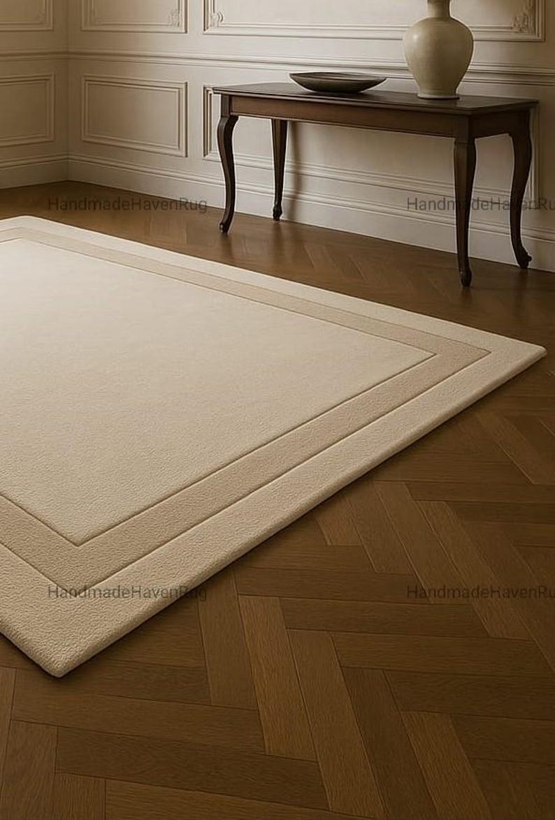 May include: A rectangular beige rug with a bordered design, placed on a herringbone pattern hardwood floor. The rug has a soft, plush texture. A dark brown table with curved legs and a decorative vase are in the background.