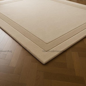May include: A rectangular beige rug with a lighter beige border, placed on a herringbone pattern wooden floor. The rug has a simple, elegant design, suitable for various interior styles. The rug is a neutral color, making it versatile for different room decors.