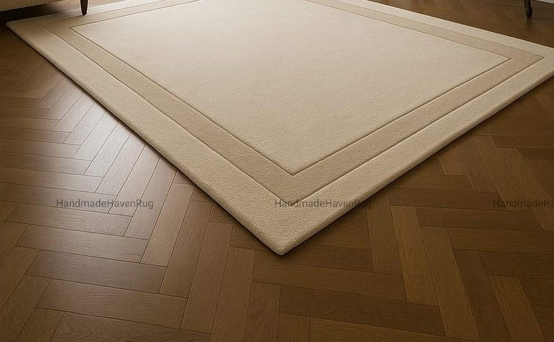 May include: A rectangular beige rug with a lighter beige border, placed on a herringbone pattern wooden floor. The rug has a simple, elegant design, suitable for various interior styles. The rug is a neutral color, making it versatile for different room decors.