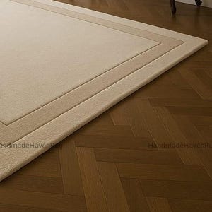 May include: A rectangular beige rug with a lighter beige border and a darker beige inner border. The rug is placed on a dark brown herringbone patterned hardwood floor. The rug's texture appears soft and plush.