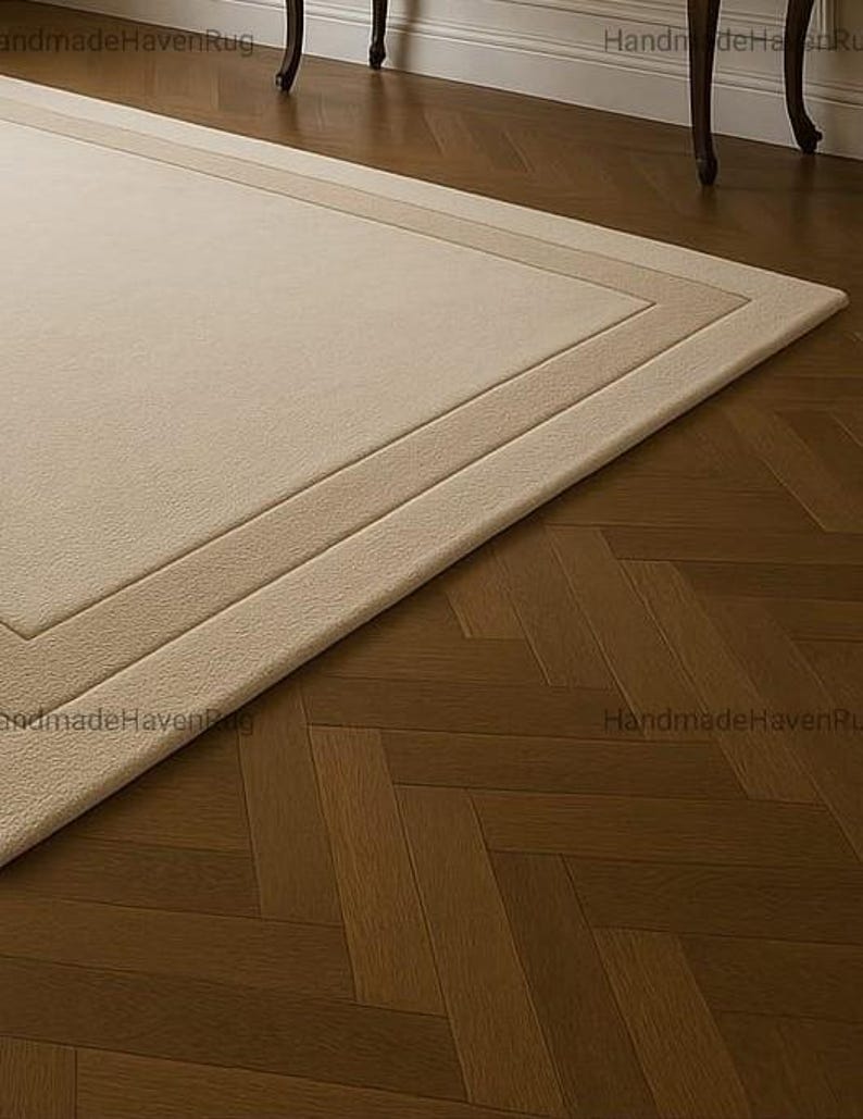 May include: A rectangular beige rug with a lighter beige border and a darker beige inner border. The rug is placed on a dark brown herringbone patterned hardwood floor. The rug's texture appears soft and plush.