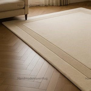 May include: Beige rectangular rug with a bordered design. The rug is placed on a wooden floor with a herringbone pattern. A beige sofa and a window with sheer curtains are in the background.