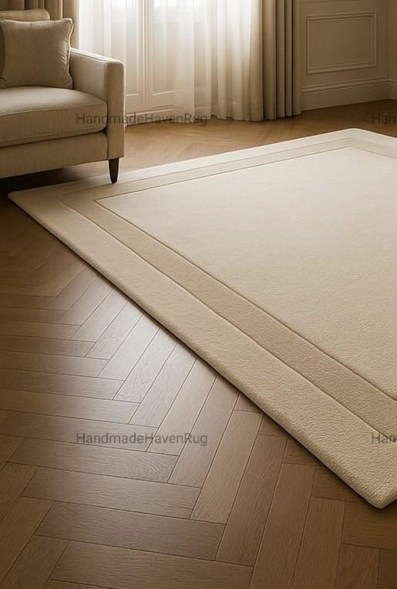 May include: Beige rectangular rug with a bordered design. The rug is placed on a wooden floor with a herringbone pattern. A beige sofa and a window with sheer curtains are in the background.