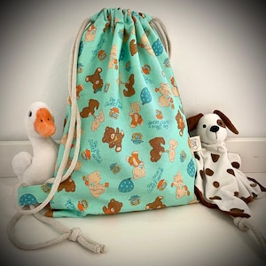 Può includere: Una borsa con coulisse verde menta con un motivo ripetuto di orsetti, vasetti di miele e testo. Un'oca bianca e un cane di peluche con macchie e orecchie marroni sono accanto alla borsa.