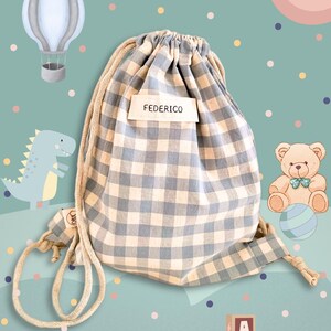 Può includere: Una borsa con coulisse a quadretti blu e bianchi con il nome "FEDERICO" su un'etichetta. La borsa è circondata da illustrazioni di una mongolfiera, un dinosauro, un orsacchiotto e blocchi dell'alfabeto, su uno sfondo turchese con pois colorati.