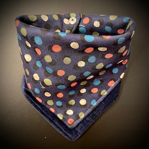 Può includere: Una bandana blu navy con un motivo a pois colorati e un bordo in velluto a coste. La bandana è piegata a forma di triangolo e fissata con una chiusura a scatto. I pois sono nelle tonalità del rosso, verde, blu e bianco.
