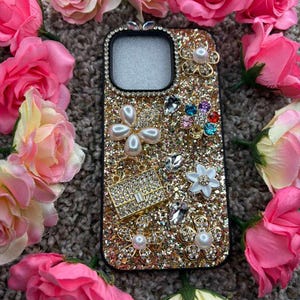 Op de afbeelding: Een met goudglitter versierde telefoonhoes, versierd met parels, strass en bloemmotieven. Omringd door roze rozen, waardoor een glamoureuze en vrouwelijke esthetiek ontstaat. Een modieus accessoire.