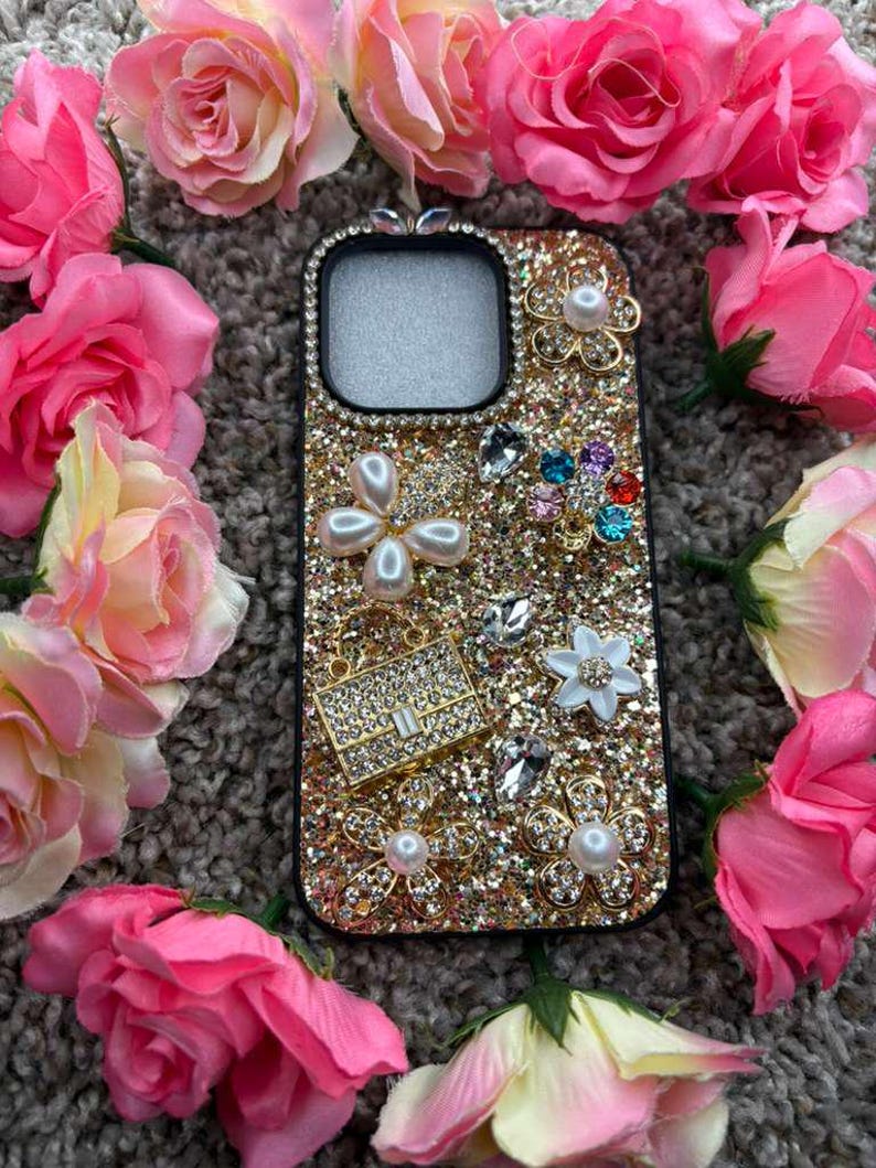Op de afbeelding: Een met goudglitter versierde telefoonhoes, versierd met parels, strass en bloemmotieven. Omringd door roze rozen, waardoor een glamoureuze en vrouwelijke esthetiek ontstaat. Een modieus accessoire.