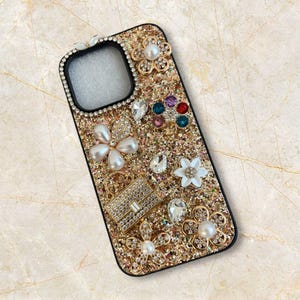 Op de afbeelding: Een met goudglitter versierde telefoonhoes, versierd met parels, strass en bloemvormige versieringen. De hoes heeft een kleine, met juwelen bezette tasjesbedel. De hoes heeft een zwarte rand rond de camera en de telefoonranden.