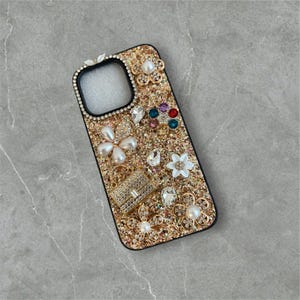 Op de afbeelding: Een gouden glitter telefoonhoesje met een zwarte rand. De hoes is versierd met parels, strass en bloemmotieven. De hoes heeft een rechthoekige vorm met een camera-uitsparing.