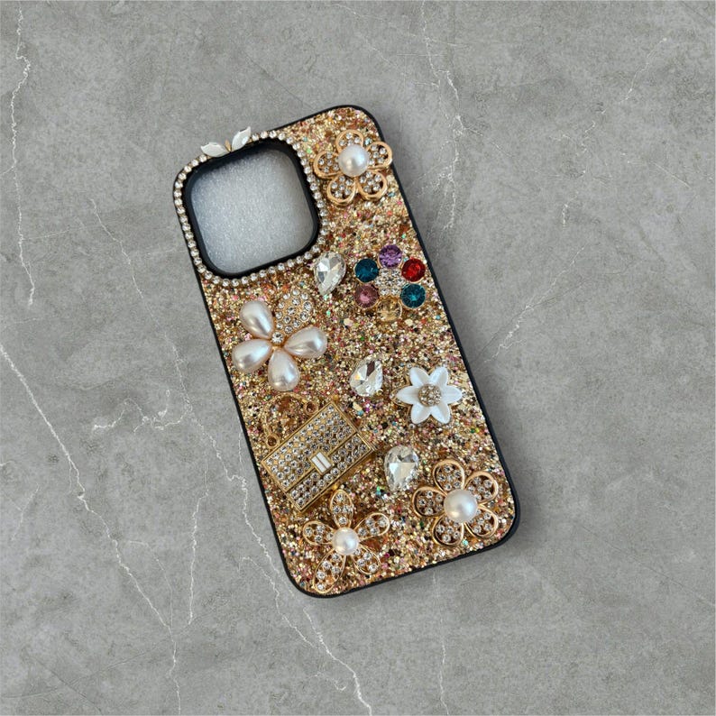 Op de afbeelding: Een gouden glitter telefoonhoesje met een zwarte rand. De hoes is versierd met parels, strass en bloemmotieven. De hoes heeft een rechthoekige vorm met een camera-uitsparing.