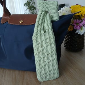 Könnte beinhalten: Hellgrüner, gehäkelter Weinflaschenhalter mit passendem Griff, der an einer marineblauen Tragetasche mit braunen Lederakzenten lehnt. Eine Vase mit bunten Blumen steht in der Nähe auf einer Holzoberfläche.
