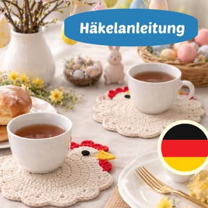 May include: A table setting featuring white teacups on cream-coloured, crocheted chicken-shaped coasters. The coasters have red, yellow, and black accents. The image includes a German flag and the text "Häkelanleitung" on a blue banner.