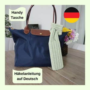 Puede incluir: Un bolso tote azul marino con detalles y asas de cuero marrón. Un artículo de ganchillo verde claro cuelga del asa. El texto en la imagen dice "Handy Tasche" y "Häkelanleitung auf Deutsch". Un ramo de flores está en el fondo.