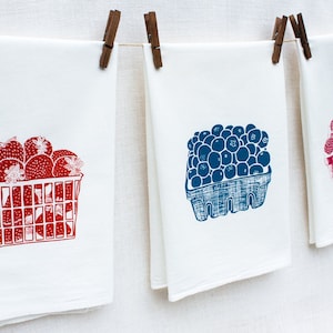 May include: Three white tea towels with red, blue, and pink printed designs of fruit in baskets. The towels are hanging on a clothesline with wooden clothespins.