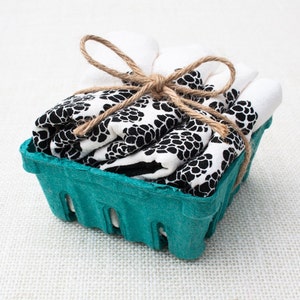 Puede incluir: Un conjunto de paños plegados con un patrón en blanco y negro, atados con hilo de bramante, presentados en una cesta de bayas verde turquesa. Los paños tienen un diseño floral negro repetido. El hilo está atado en un lazo.