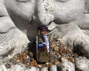 May include: A small clear glass bottle with a cork stopper containing dried herbs, a white ribbon, and a blue and white evil eye bead. The bottle is positioned upright, filled with green botanical matter and a small rolled paper scroll inside.