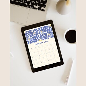 May include: A digital tablet shows a January 2026 calendar with a blue and yellow floral pattern. The tablet sits on a white surface, alongside a laptop, a coffee cup, and a pen. The calendar has a cream background.