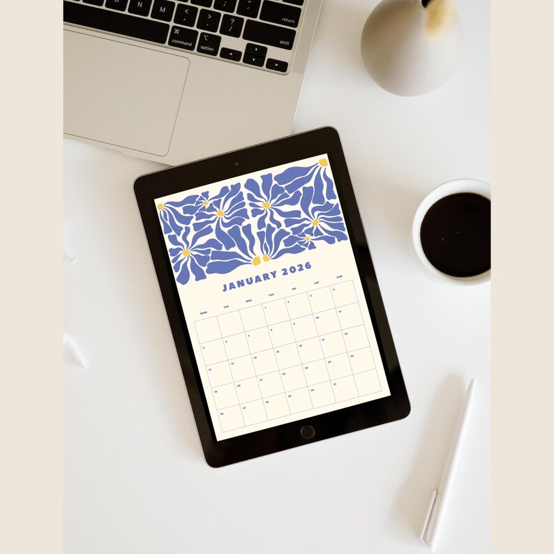 May include: A digital tablet shows a January 2026 calendar with a blue and yellow floral pattern. The tablet sits on a white surface, alongside a laptop, a coffee cup, and a pen. The calendar has a cream background.