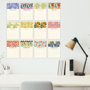 May include: Twelve monthly calendars with cream-coloured backgrounds and vibrant floral designs. The calendars are displayed on a white wall above a desk with a lamp, books, and a plant, creating a decorative display.