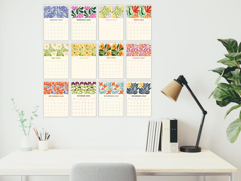 May include: Twelve monthly calendars with cream-coloured backgrounds and vibrant floral designs. The calendars are displayed on a white wall above a desk with a lamp, books, and a plant, creating a decorative display.