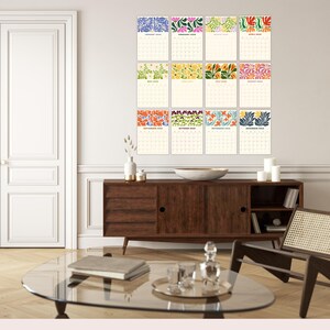 May include: A collection of twelve calendars, each featuring a floral design in various colours. The calendars are displayed on a white wall above a dark wood cabinet. Each calendar is labelled with a month of the year.