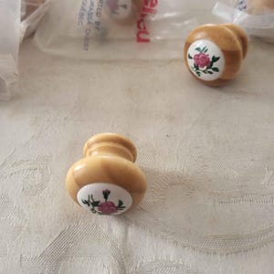 Puede incluir: Dos pomos redondos de madera para armarios con acabado marrón claro y una incrustación circular blanca en el centro. Cada incrustación presenta un pequeño gráfico de una rosa rosa con hojas verdes. Un pomo está suelto, otros en plástico.
