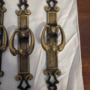Op de afbeelding: Close-up van verschillende antieke messing deurgrepen. Elke greep heeft een decoratieve handgreep met een gebogen ontwerp, een rechthoekig midden met een houtnerftextuur en sierlijke boven- en onderdetails. De grepen zijn gerangschikt op een witte doek.