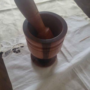 Könnte beinhalten: Ein Mörser mit Stößel aus Holz, bestehend aus einer zylindrischen Schale mit verbreitertem Fuß und einem passenden abgerundeten Stößel. Das Holz zeigt eine zweifarbige Maserung in Hell- und Dunkelbraun sowie einen sichtbaren vertikalen Riss.