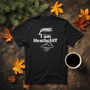 May include: Black t-shirt featuring the white text "I am Heathcliff", a tree graphic, and a storm cloud illustration. The shirt is set against a backdrop of autumn leaves and foliage, with a coffee cup in the bottom right.