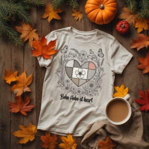 Könnte beinhalten: Cremefarbenes T-Shirt mit herzförmigem Patchwork-Design, umgeben von floralen und Schmetterlingsillustrationen. Der Text "Boho Hobo at heart" ist darunter gedruckt. Das Bild ist auf einer Holzoberfläche mit Herbstblättern, einem Kürbis und einer Tasse Kaffee platziert.