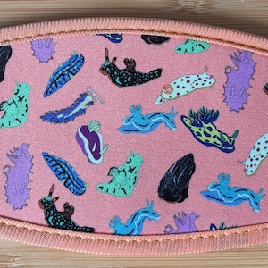 May include: A coral-colored face mask featuring a repeating pattern of colorful sea slugs in various shapes and colors. The mask has a peach-colored trim and is made of fabric.