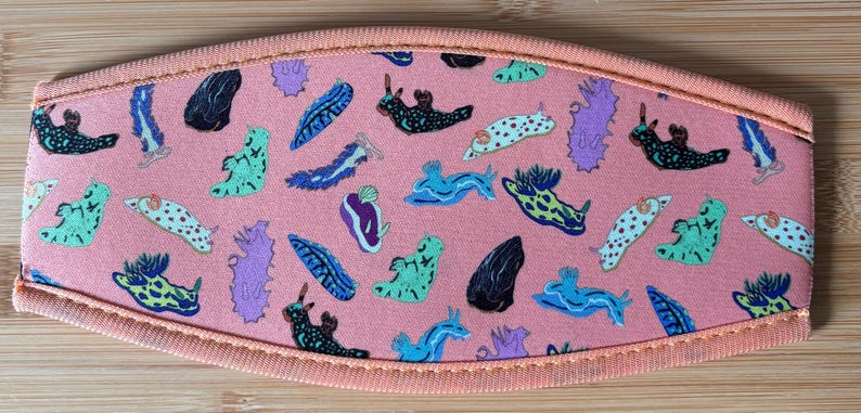 May include: A coral-colored face mask featuring a repeating pattern of colorful sea slugs in various shapes and colors. The mask has a peach-colored trim and is made of fabric.