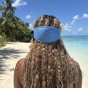 May include: A light blue headband with a white dolphin pattern is worn on a person's head. The person is on a sandy beach with palm trees and a clear blue sky in the background. The headband is made of a soft material.