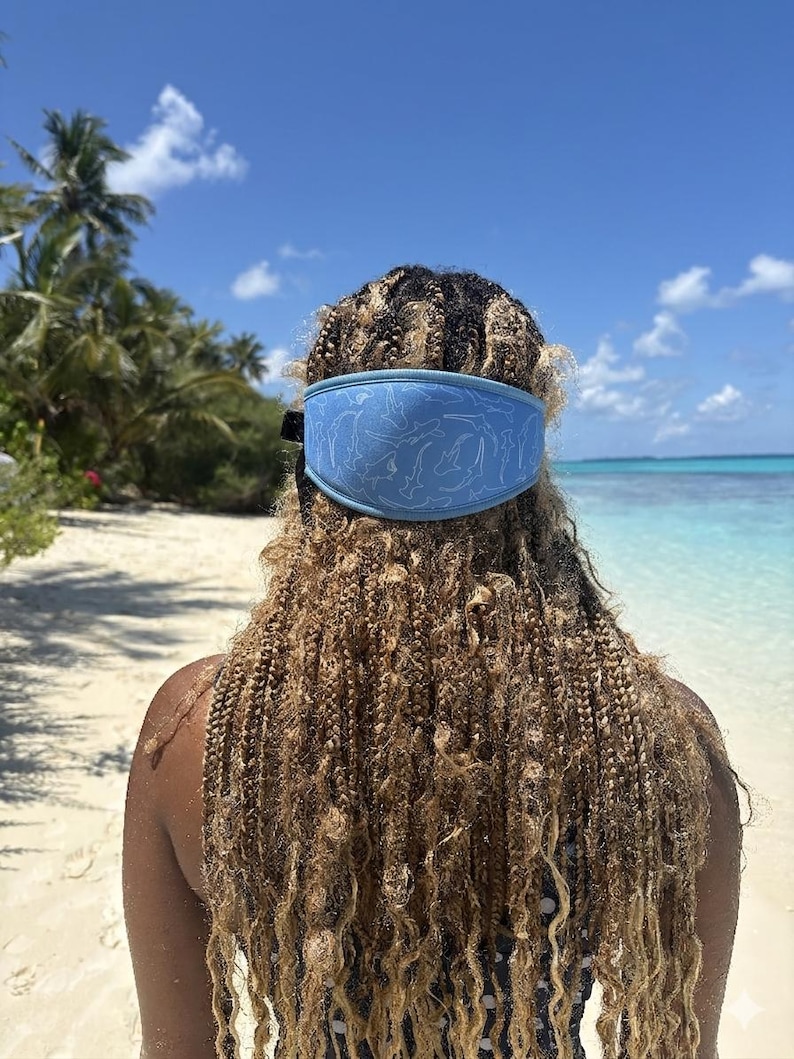 May include: A light blue headband with a white dolphin pattern is worn on a person's head. The person is on a sandy beach with palm trees and a clear blue sky in the background. The headband is made of a soft material.
