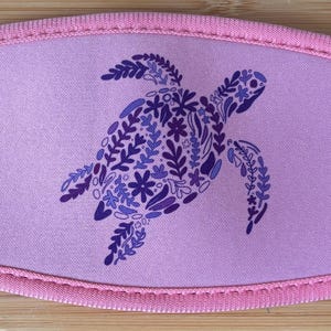 May include: A lavender face mask featuring a sea turtle design. The turtle is composed of blue and purple floral patterns. The mask has a pink trim and is made of a soft, flexible material.