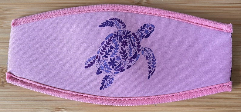 May include: A lavender face mask featuring a sea turtle design. The turtle is composed of blue and purple floral patterns. The mask has a pink trim and is made of a soft, flexible material.