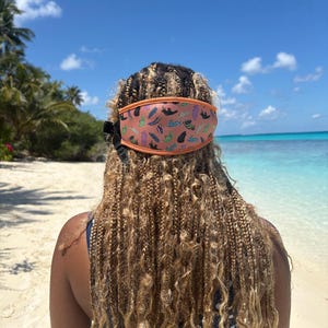 May include: A coral-colored face mask with an orange trim and a pattern of various shapes and colors. The mask is worn on a person's head, with their braided hair cascading down their back. The background features a beach, ocean, and blue sky.