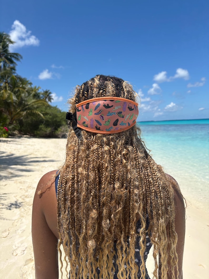 May include: A coral-colored face mask with an orange trim and a pattern of various shapes and colors. The mask is worn on a person's head, with their braided hair cascading down their back. The background features a beach, ocean, and blue sky.