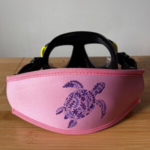 May include: A black and yellow scuba diving mask with a pink strap cover. The strap cover features a purple floral sea turtle design. The mask is set on a wooden surface.
