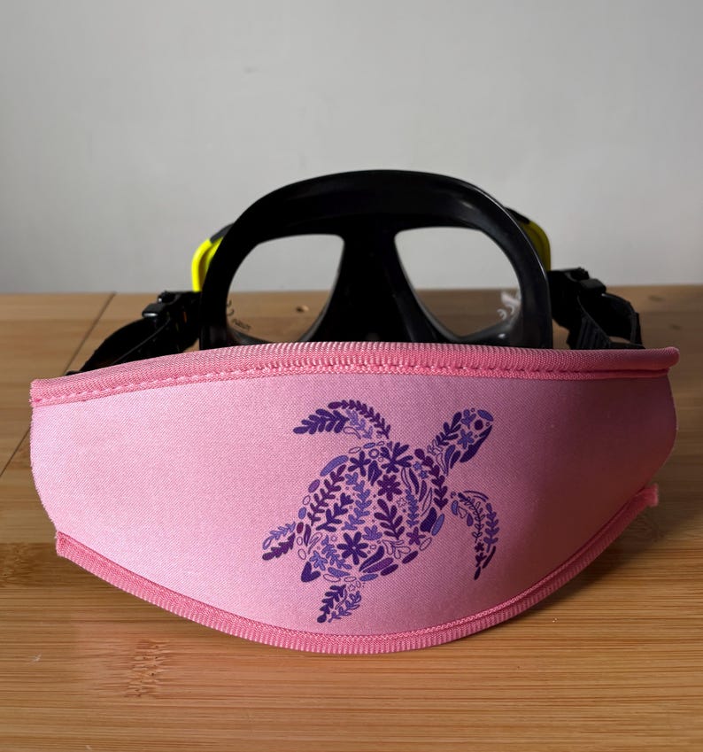 May include: A black and yellow scuba diving mask with a pink strap cover. The strap cover features a purple floral sea turtle design. The mask is set on a wooden surface.