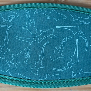 May include: Teal-colored beverage insulator with a white shark outline pattern. The edges are trimmed with a matching teal fabric and stitching. The design features various shark species.