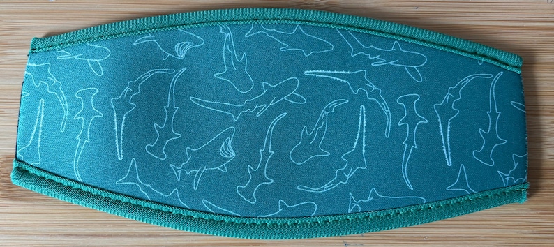 May include: Teal-colored beverage insulator with a white shark outline pattern. The edges are trimmed with a matching teal fabric and stitching. The design features various shark species.