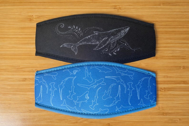 May include: Two face masks. The top mask is black with a white whale design. The bottom mask is blue with a white shark pattern. Both masks have a curved shape and are made of a soft material.