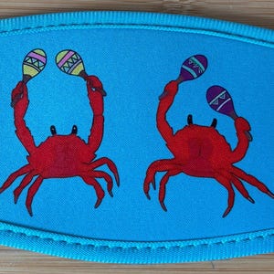 May include: A light blue drink sleeve featuring two red crabs holding maracas. The maracas have yellow, purple, and green accents. The sleeve has a stitched border.