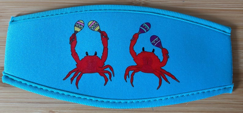 May include: A light blue drink sleeve featuring two red crabs holding maracas. The maracas have yellow, purple, and green accents. The sleeve has a stitched border.