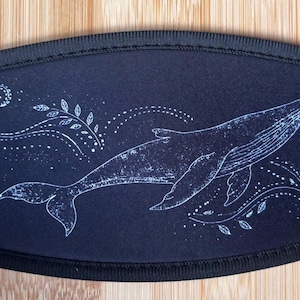 May include: A black face mask featuring a white illustration of a whale swimming. The whale is surrounded by decorative swirls and dotted lines. The mask has a black border and is set against a wooden background.