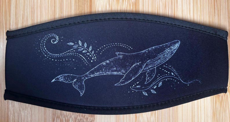 May include: A black face mask featuring a white illustration of a whale swimming. The whale is surrounded by decorative swirls and dotted lines. The mask has a black border and is set against a wooden background.