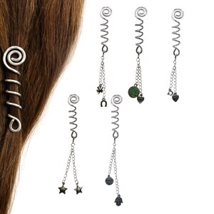 Op de afbeelding: Een set van vijf zilverkleurige metalen haarspiralen, elk met een spiraalvormige bovenkant en een kettinkje met diverse bedels, waaronder een klavertjevier, hoefijzer, boom, hartje, sterren, een ronde schijf en een hamsa-hand symbool.