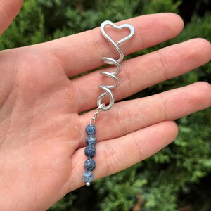 Op de afbeelding: Een zilverkleurige metalen haarspiraal met een hartvormige bovenkant en een bungelende bedel bestaande uit vijf kleine, ronde, getextureerde blauwe kralen. De spiraal is gemaakt van een enkele draad die naar beneden krult vanaf het hart.